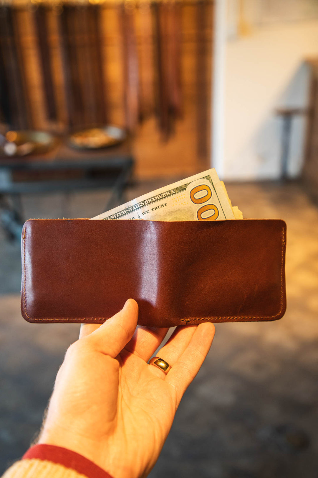 Hand holding a brown leather wallet with $100 bills against a blurred indoor background #color_brown