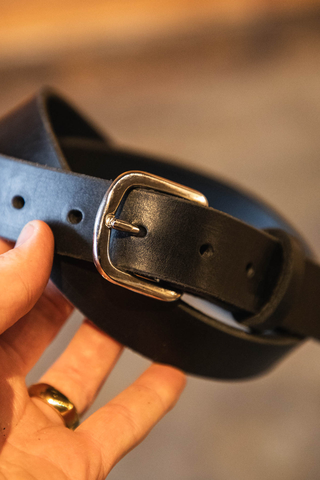Black leather belt with silver buckle held by a hand against a blurred background #color_black