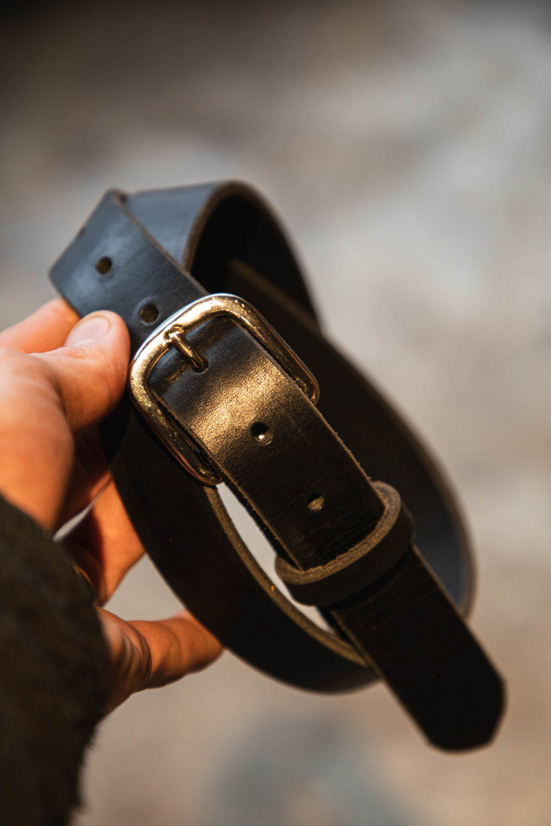 Black leather belt with nickel buckle held by a hand against a blurred background #color_black