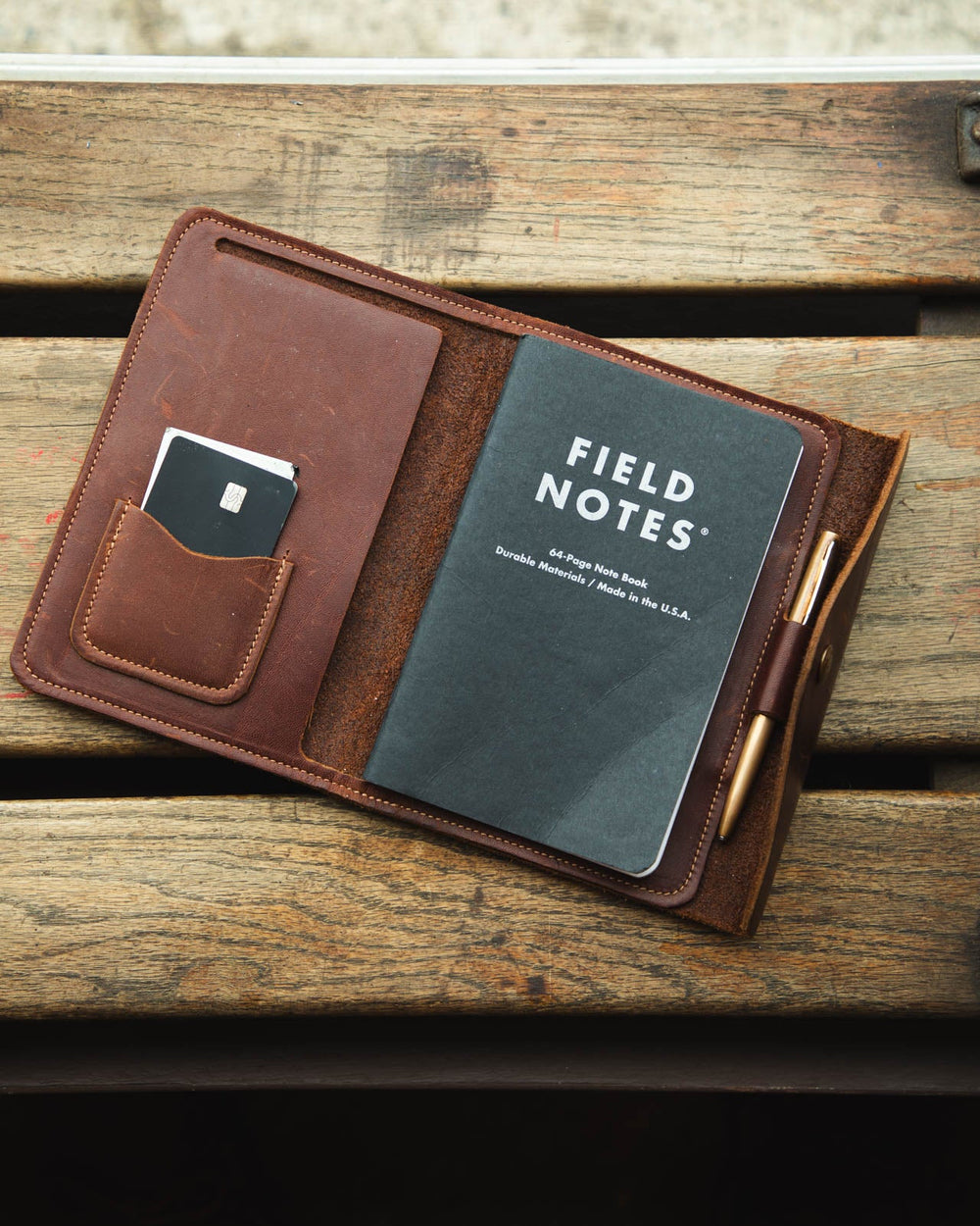 A brown leather organizer case laid open on a rustic wooden surface, holding a dark Field Notes notebook, a card in a side pocket, and a pen loop with a gold pen.