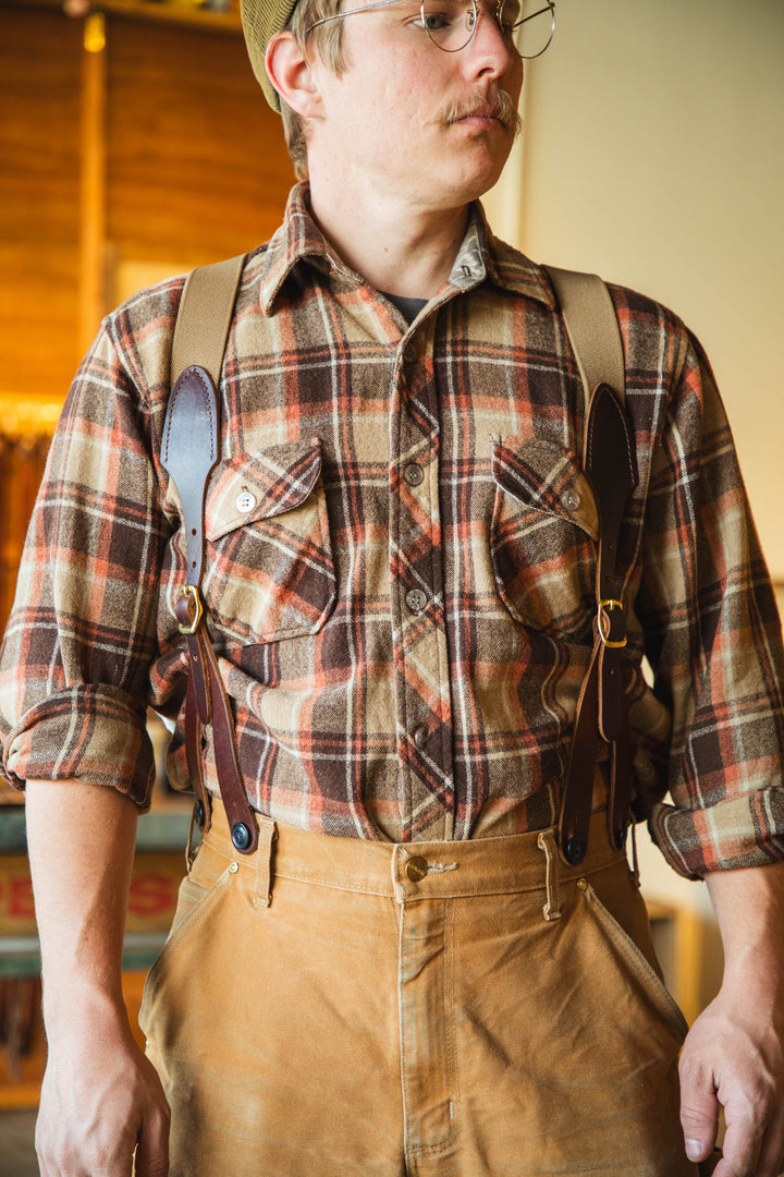 Man wearing a plaid shirt, suspenders, and overalls in an indoor setting.