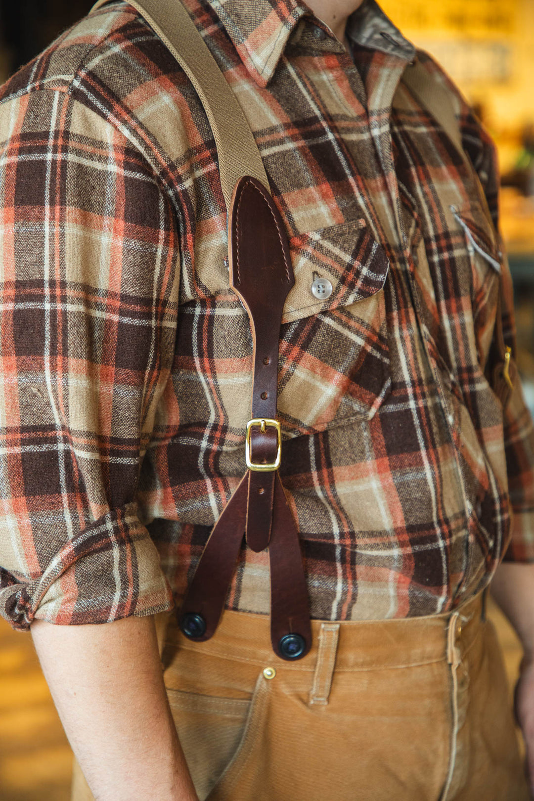 Person wearing a plaid shirt with suspenders and brown pants against a blurred background
