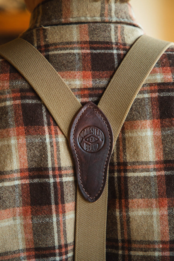 Close-up of a plaid shirt with suspenders and a leather strap featuring &